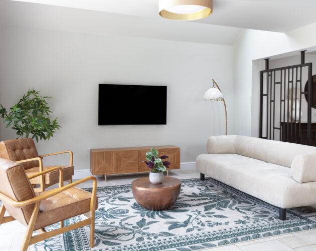 Open plan lounge area with stylish sofas in cream and tan leather. plants. wall mounted tV with walnut and gold sideboard below. Round flush ceiling light. japanese inspired floor lamp. Black metal room divider with dark stained wood cabinet behind.