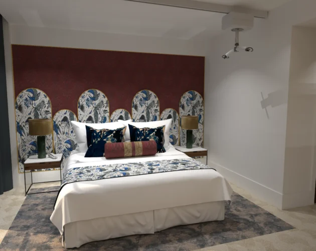 A stylish, disability-friendly hotel bedroom with a ceiling hoist above the bed. The bed features a multi-panel floral and bird-patterned headboard against a red accent wall, dressed in white linens with blue, gold, and red pillows. Wooden bedside tables with green lamps and a patterned rug complete the elegant, accessible design.