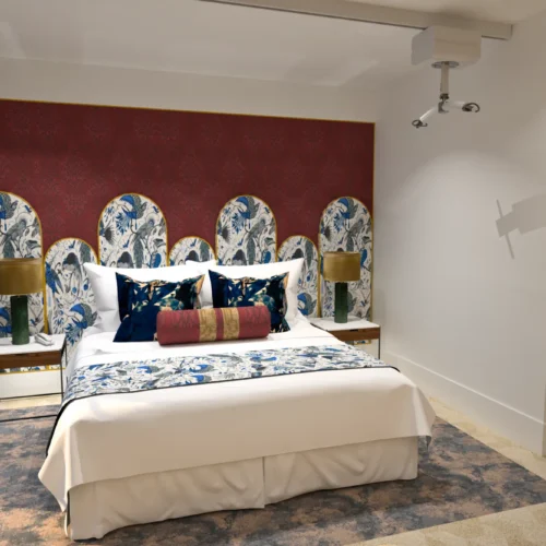 A stylish, disability-friendly hotel bedroom with a ceiling hoist above the bed. The bed features a multi-panel floral and bird-patterned headboard against a red accent wall, dressed in white linens with blue, gold, and red pillows. Wooden bedside tables with green lamps and a patterned rug complete the elegant, accessible design.