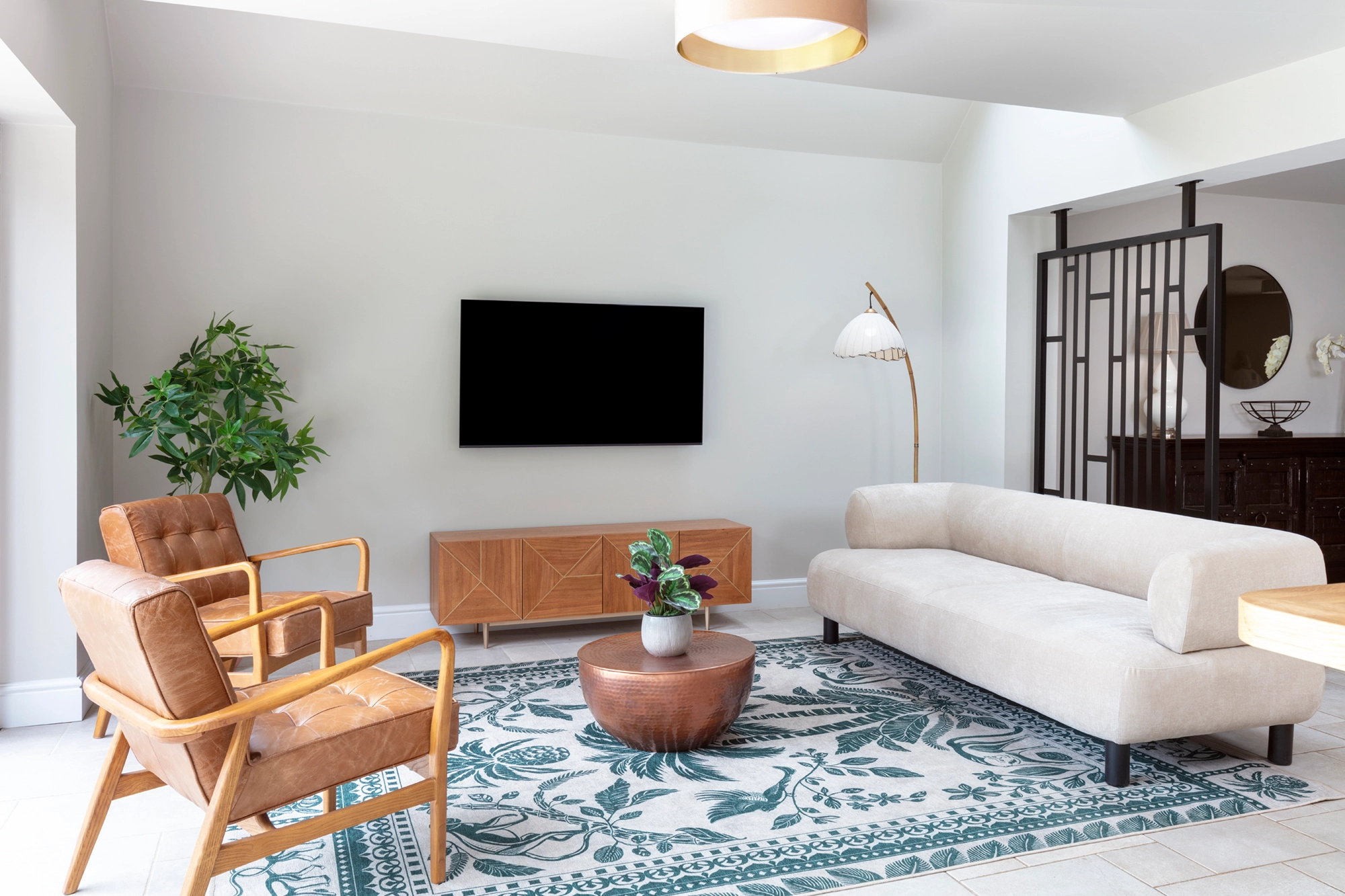 Open plan lounge area with stylish sofas in cream and tan leather. plants. wall mounted tV with walnut and gold sideboard below. Round flush ceiling light. japanese inspired floor lamp. Black metal room divider with dark stained wood cabinet behind.